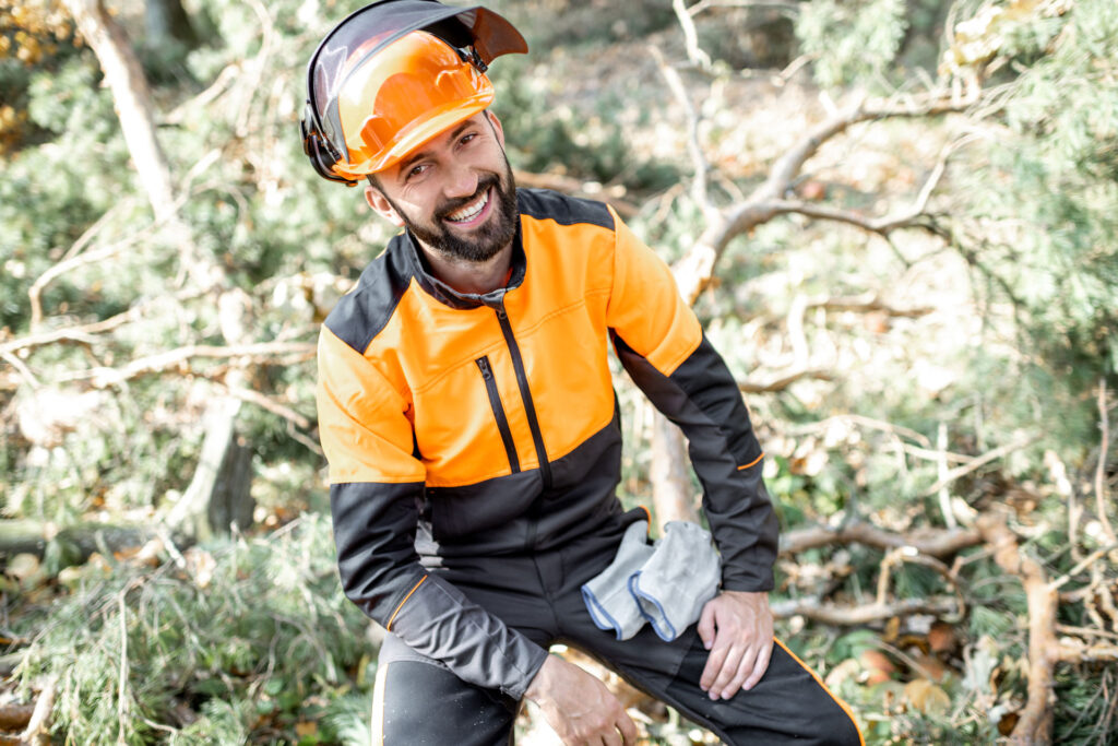 Forstarbeiter, Holzarbeiter in professioneller Schutzkleidung und Helm – Portrait in der Pause. im Wald. Sympatischer Mann zwischen 35 und 45 Jahren mit Vollbart, lächelnd